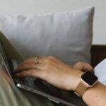 Smart Home Sensors - Side view of crop unrecognizable female freelancer in casual clothes and smartwatch lying on comfortable sofa and working remotely on laptop